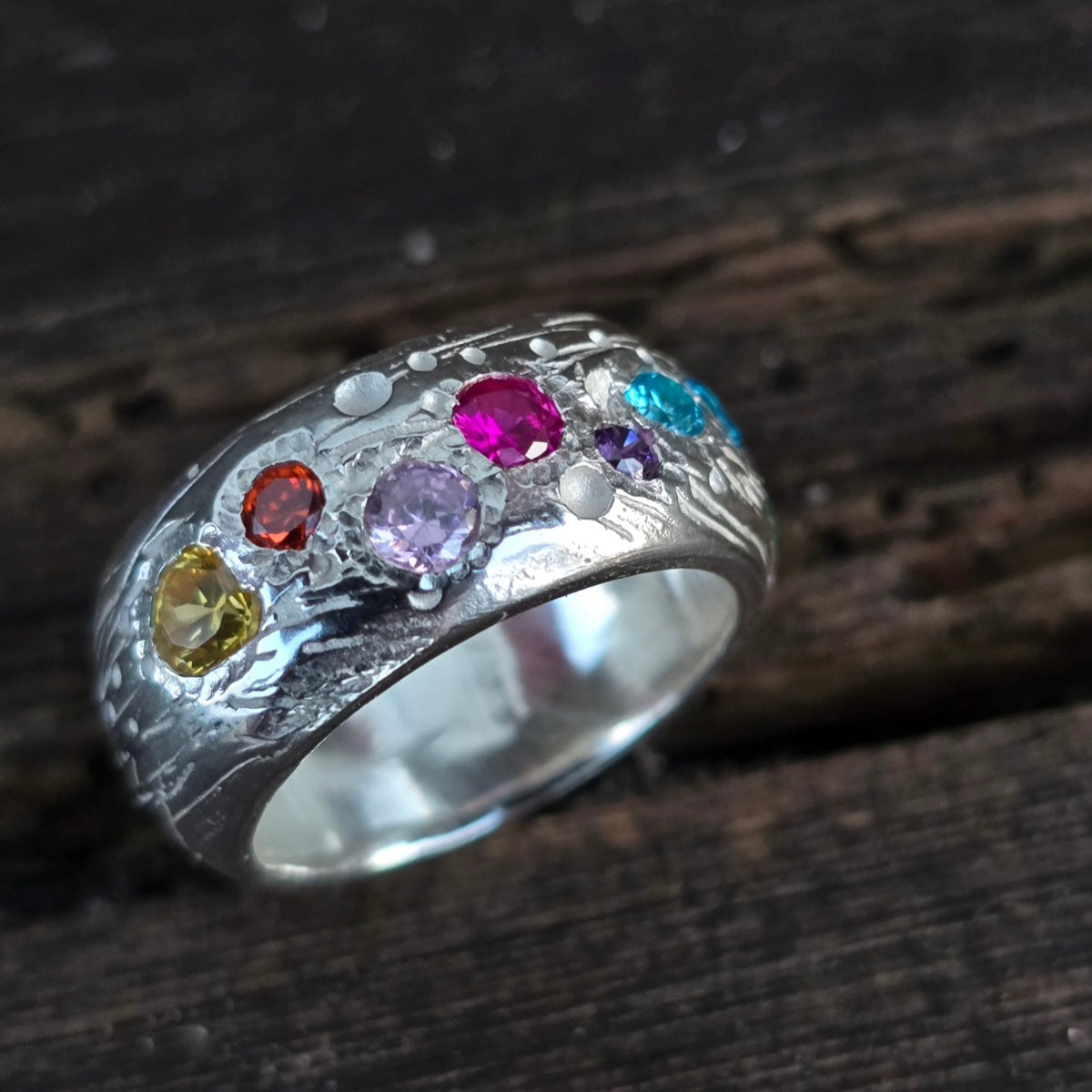 Breiter handgefertigter Silberring mit bunter Edelsteinbesatzung in Blau, Pink, Violett, Orange und Gelb, auf dunklem Holz fotografiert. Unikatschmuck mit Strukturmuster.