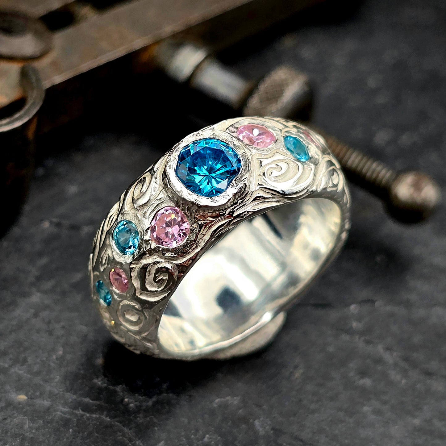 Ein massiver, handgefertigter Silberring liegt auf dunklem Untergrund. Der Ring ist breit und mit Schnörkelmustern verziert. Mehrere runde Edelsteine in leuchtendem Blau und zartem Rosa sind kunstvoll in die Oberfläche eingefasst. Im Mittelpunkt steht ein großer, funkelnder blauer Edelstein.