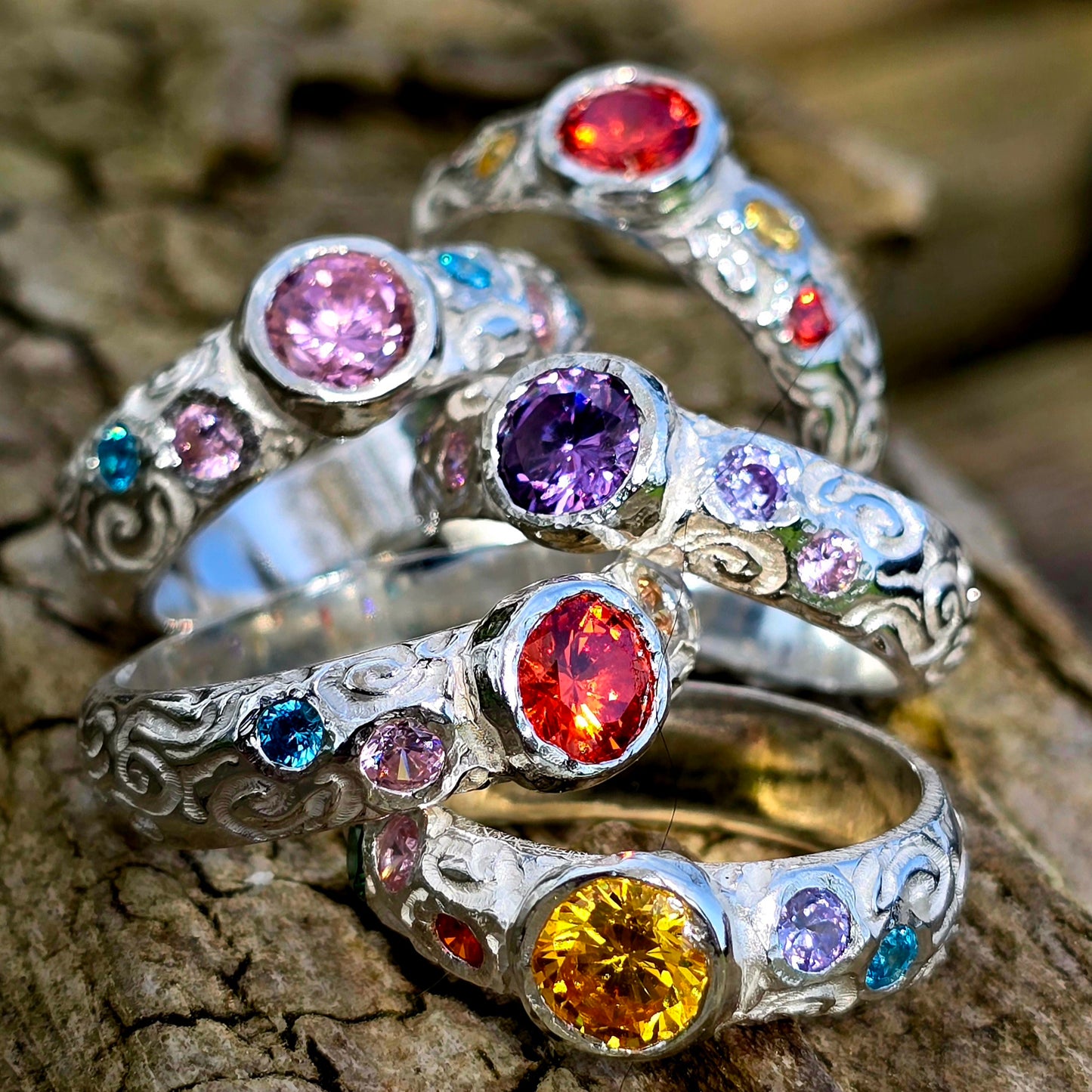 Fünf handgefertigte Silberringe liegen übereinander auf einem Stück Holz. Jeder Ring trägt mehrere bunte Edelsteine in kräftigen Farben wie Rot, Gelb, Lila, Rosa und Blau. Die Ringe sind mit feinen Schnörkeln und Mustern verziert und funkeln im Sonnenlicht. Sie erinnern an die Farben eines Regenbogens.