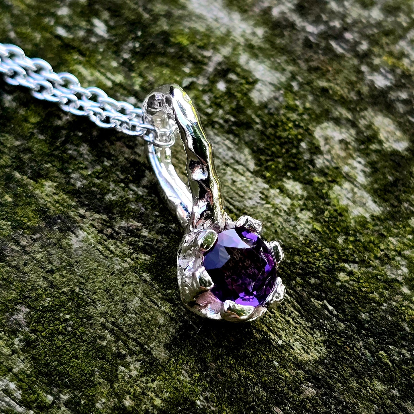 Handgefertigter Anhänger aus Silber mit einem runden, facettierten Amethyst in leuchtendem Violett. Der tief violette Edelstein ist von einer organischen, leicht geschwungenen Fassung umgeben, die an florale Formen erinnert. Einzelstück mit strukturierter Oberfläche, modern und naturinspiriert – perfekt als Schmuckgeschenk oder für Liebhaber von Edelsteinschmuck.