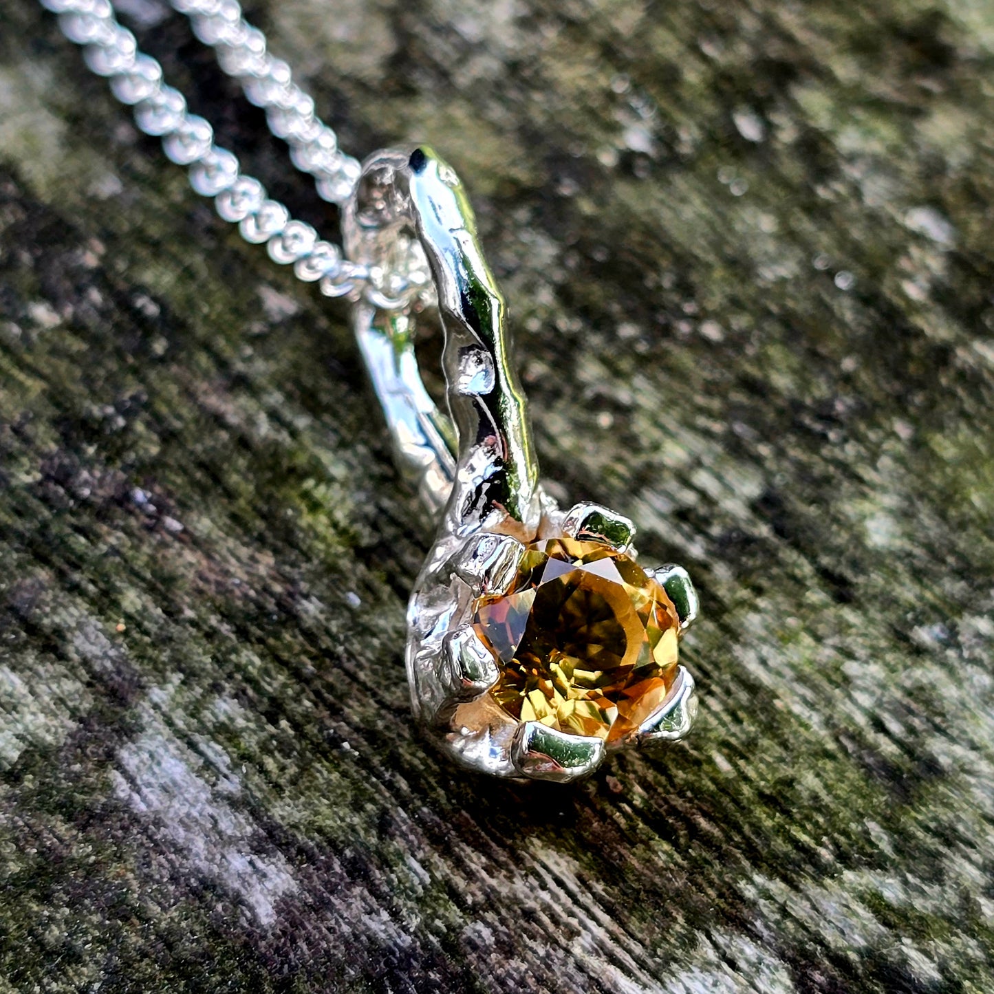 Nahaufnahme eines handgefertigten Silberanhängers mit gelbem Edelstein. Der Anhänger zeigt ein organisches Design in floraler oder krallenartiger Form und umfasst den facettierten Edelstein wie eine schützende Hülle. Der Schmuck liegt auf verwittertem Holz mit Moosstruktur. Moderner Unikatschmuck aus Silber – ideal für Natur- und Fantasy-Liebhaber*innen.