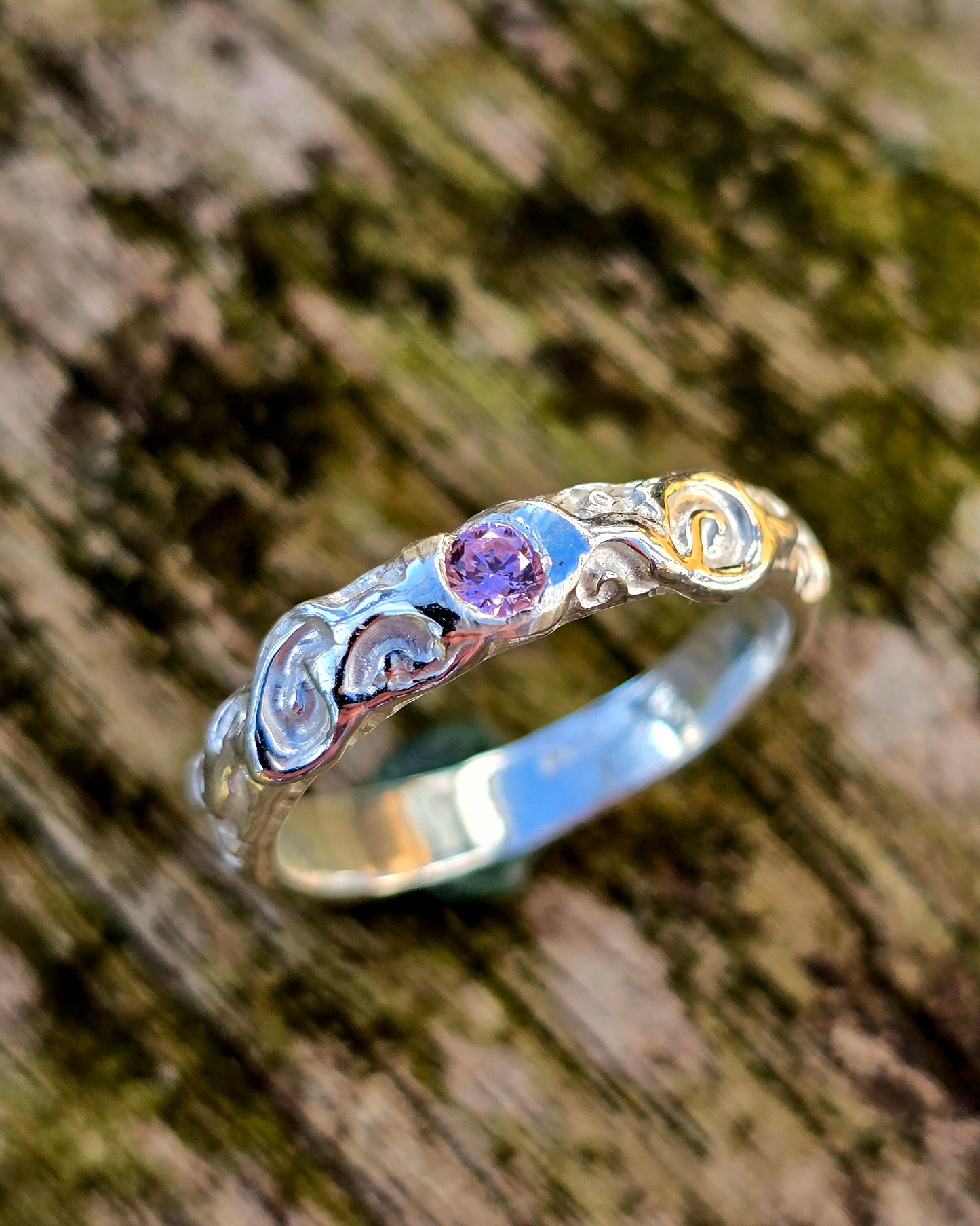 Handgefertigter Silberring mit zarten, verschnörkelten Details und einem funkelnden rosa Saphir. Ein kunstvolles Unikat im Lost-Wax-Verfahren gefertigt, präsentiert auf rustikalem Holz.
