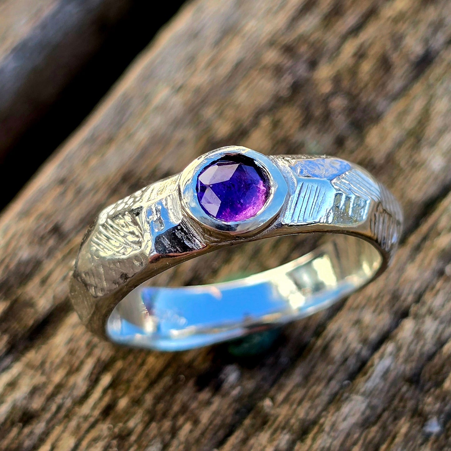 Handgefertigter Silberring mit facettiertem Amethyst, verziert mit einzigartigen Gravurmustern. Ein kunstvolles Unikat aus dem Lost-Wax-Verfahren, präsentiert auf rustikalem Holz