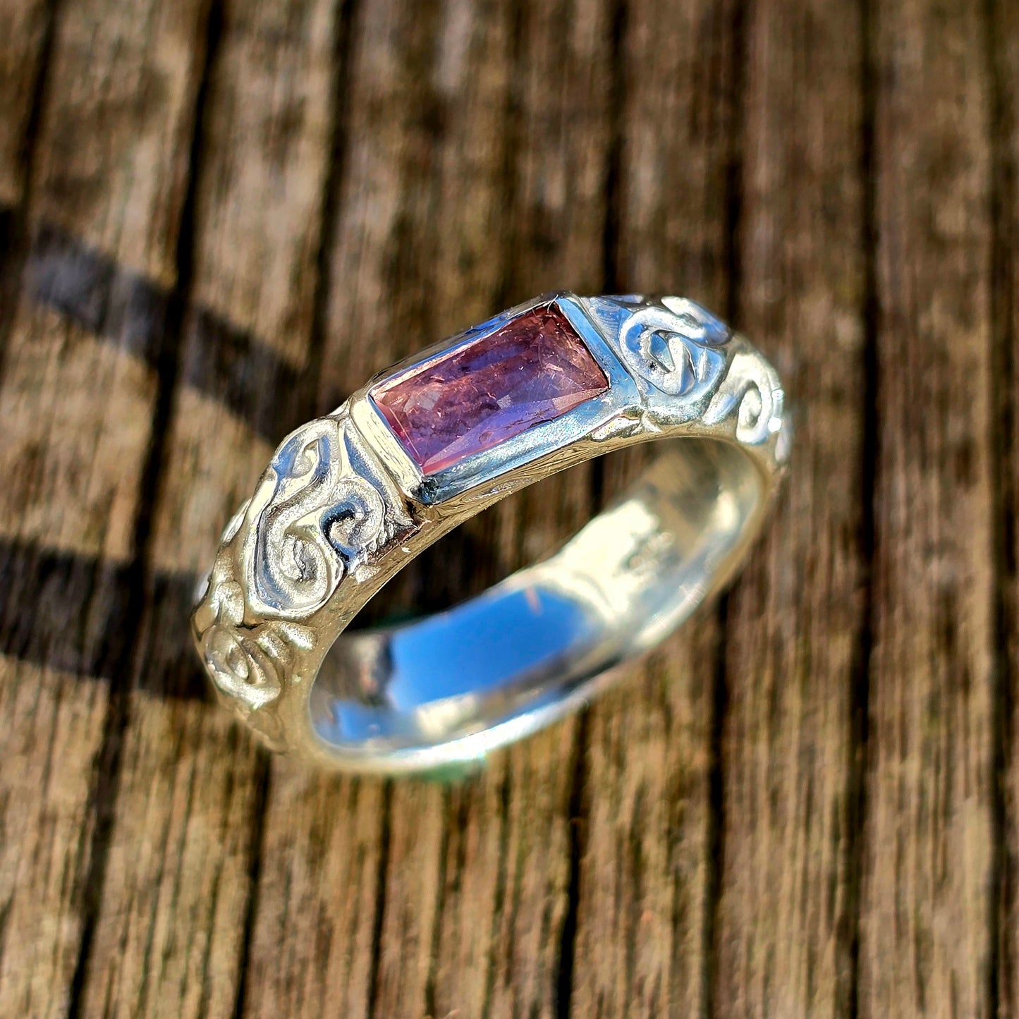Handgefertigter Silberring mit rechteckigem rosa Turmalin im Rosenschliff. Der Edelstein ist quer gefasst und schimmert in zarten Rosatönen. Die breite Ringschiene ist detailreich mit floralen Schnörkeln verziert und verleiht dem Ring eine verspielte, romantische Ausstrahlung. Einzelstück aus der Schnörkel-Kollektion.