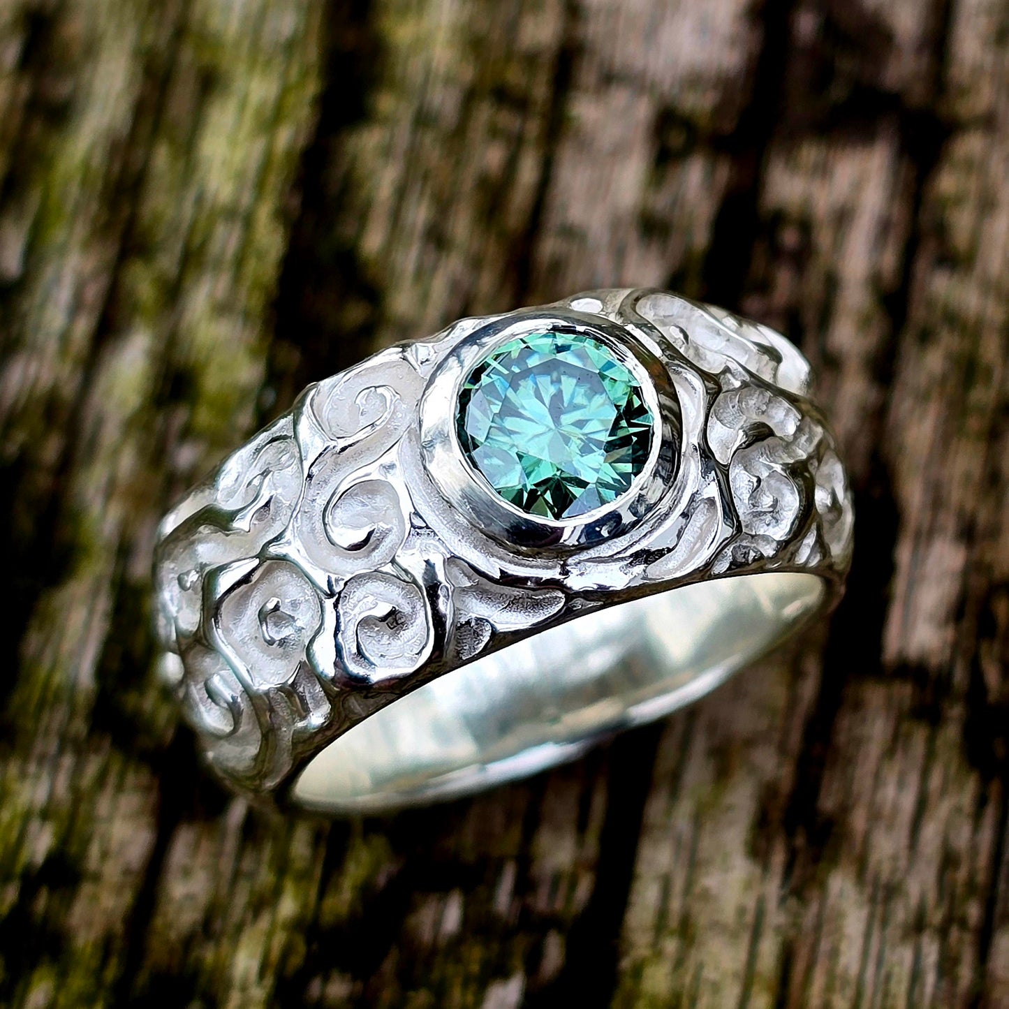 Silberring mit mittig gefasstem, rundem Moissanit in mintgrünem Farbton. Der Edelstein ist brillant geschliffen und schimmert je nach Lichteinfall grünlich-bläulich. Die Ringschiene ist breit und komplett von Hand mit verspielten Schnörkeln und stilisierten Rosenstrukturen verziert. Der Ring stammt aus der Schnörkel-Kollektion und ist ein Einzelstück aus recyceltem Silber – ideal als Statement-Ring oder besonderer Verlobungsring