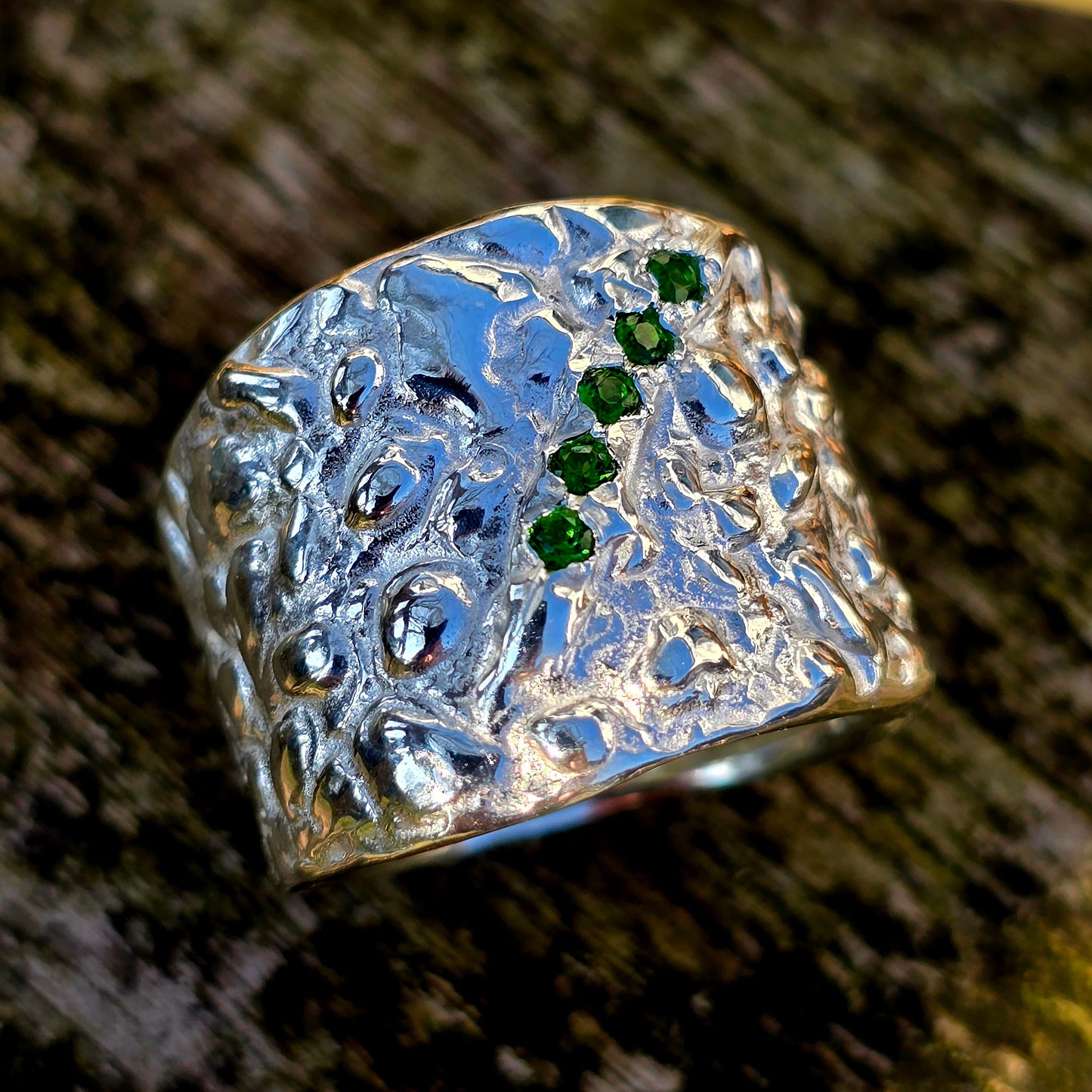 Silberring mit organischer, strukturierter Avocado Oberfläche und fünf funkelnden grünen Chromdiopsid-Edelsteinen in einer diagonalen Anordnung. Der breite Ring hat ein handgefertigtes, rustikales Design und liegt auf einem hölzernen Untergrund. Ideal für Liebhaber einzigartiger, kunstvoller Schmuckstücke.