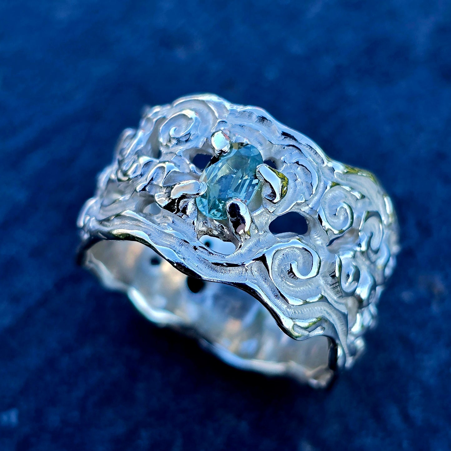 Handgefertigter Silberring mit Natur-Saphir, Einzelstück in Größe 59. Der Ring zeigt eine breite, kunstvoll verzierte Ringschiene mit floralen Schnörkeln, eingefasst ist ein heller, ovaler Saphir.
