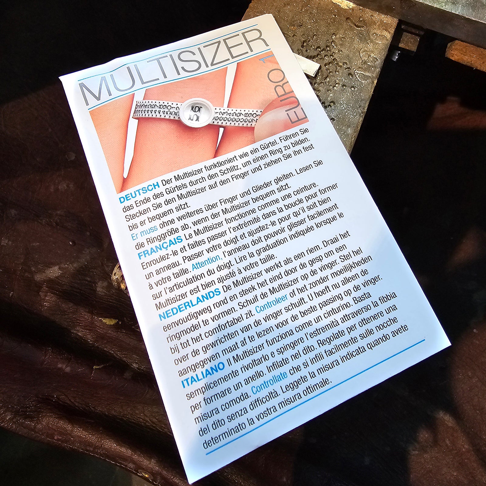 Ringgrößenmesser Multisizer aus Kunststoff, auf einem Tisch aus dunklem Holz fotografiert. Hilfsmittel zur Bestimmung der Ringgröße, Anleitung in mehreren Sprachen.
