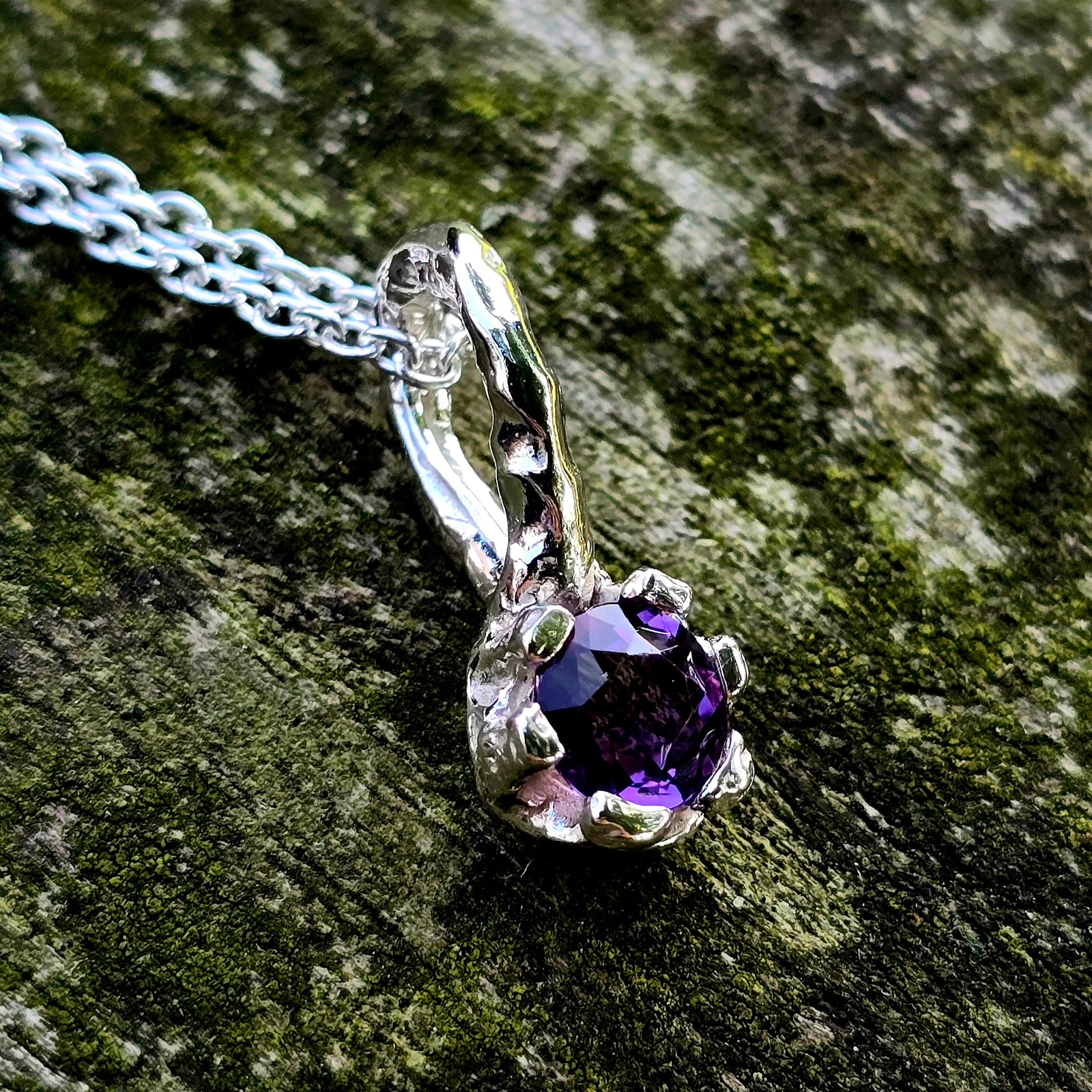 Handgefertigter Anhänger aus Silber mit einem runden, facettierten Amethyst in leuchtendem Violett. Der tief violette Edelstein ist von einer organischen, leicht geschwungenen Fassung umgeben, die an florale Formen erinnert. Einzelstück mit strukturierter Oberfläche, modern und naturinspiriert – perfekt als Schmuckgeschenk oder für Liebhaber von Edelsteinschmuck.