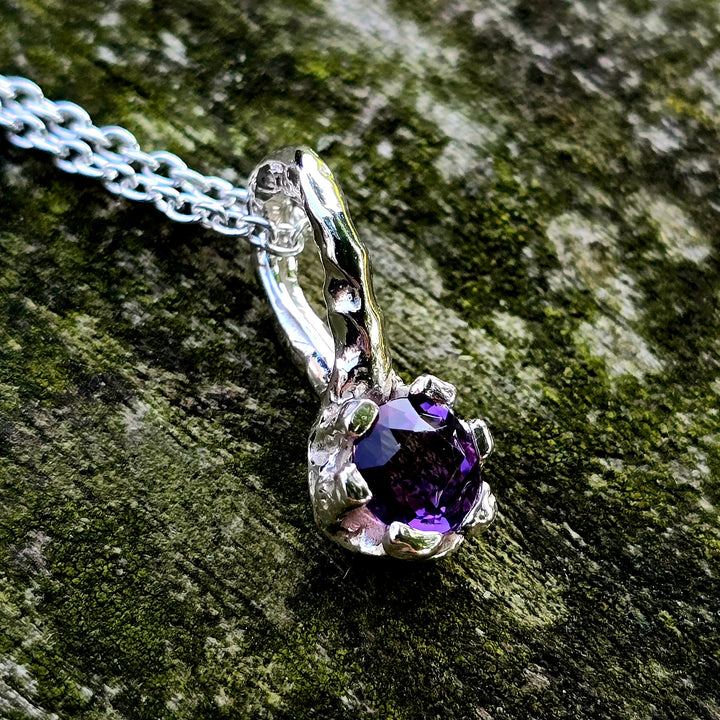 Handgefertigter Anhänger aus Silber mit einem runden, facettierten Amethyst in leuchtendem Violett. Der tief violette Edelstein ist von einer organischen, leicht geschwungenen Fassung umgeben, die an florale Formen erinnert. Einzelstück mit strukturierter Oberfläche, modern und naturinspiriert – perfekt als Schmuckgeschenk oder für Liebhaber von Edelsteinschmuck.