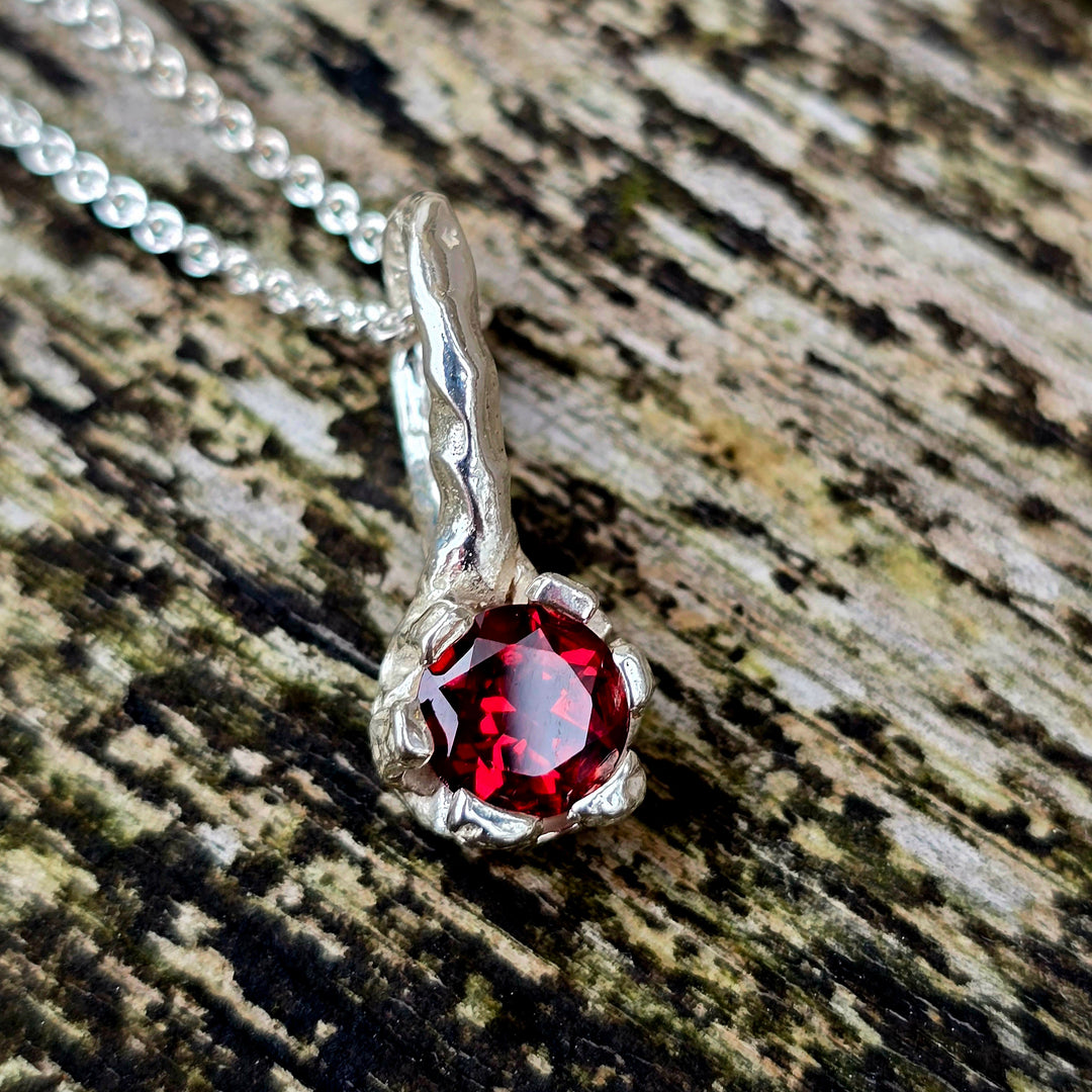 Granat-Anhänger aus Silber, handgefertigt mit organischer Krappenfassung. Der runde rote Edelstein ist ein Granat – Geburtsstein des Monats Januar. Die geschwungene Öse erinnert an Naturformen. Unikat-Schmuck für Liebhaber naturinspirierter Designs und ausdrucksstarker Edelsteine.