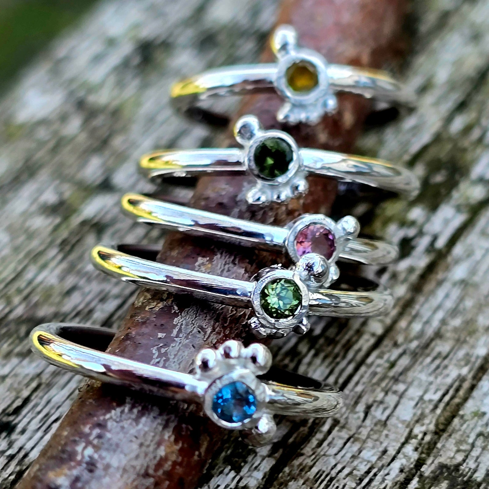 Fünf handgeschmiedete, zarte Silberringe aus der Dainty Kollektion, jeder besetzt mit einem kleinen farbigen Edelstein – gelber Saphir, dunkelgrüner Saphir, rosa Saphir, grüner Peridot und blauer Topas – auf einem rustikalen Holzast fotografiert