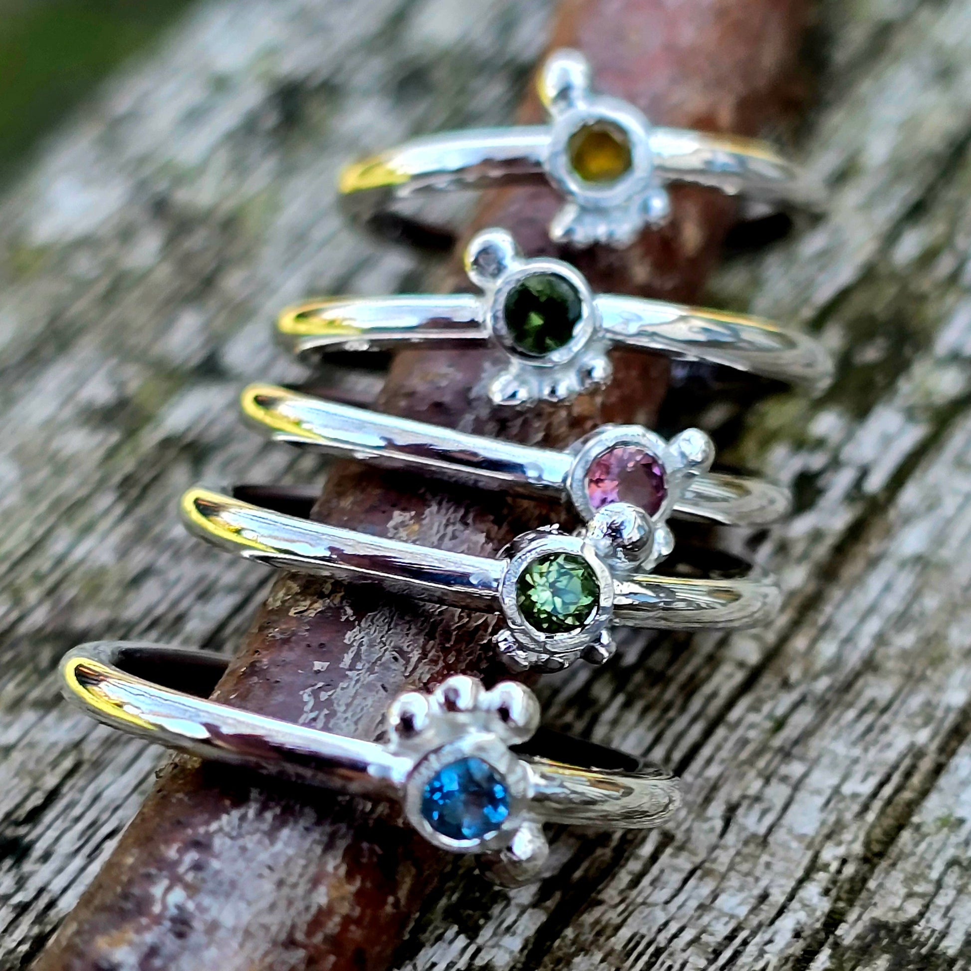 Fünf handgeschmiedete, zarte Silberringe aus der Dainty Kollektion, jeder besetzt mit einem kleinen farbigen Edelstein – gelber Saphir, dunkelgrüner Saphir, rosa Saphir, grüner Peridot und blauer Topas – auf einem rustikalen Holzast fotografiert